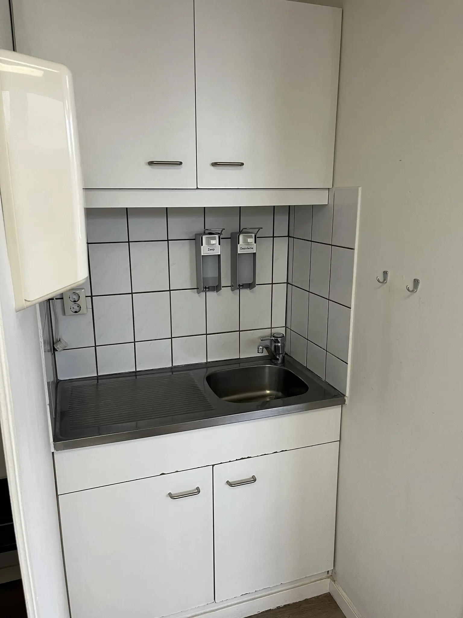 Kleine kitchenette met wit tegelwandje, roestvrijstalen spoelbak en zeepdispensers aan de Burgemeester Smitsstraat.